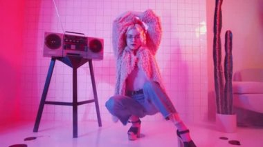 Göz kamaştırıcı kıyafetli genç sarışın kadın dansçı Vogue elleri gösterisi ve kamera izlerken stüdyoda pembe neon ışıklı retro kaset çaların yanında oturuyor.