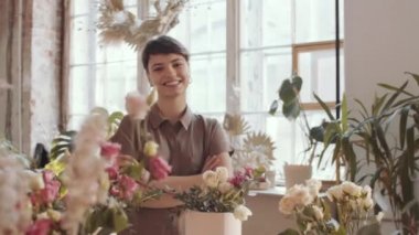 Kolları çaprazlanmış güzel güllerin arkasında durup çiçekçide poz verirken kameraya bakıp mutlu bir şekilde gülümseyen kısa saçlı neşeli kadın çiçekçinin kemer kemeri.