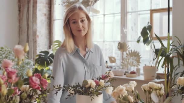 Arc shot of beautiful blonde female florist arranging roses composition in hatbox and then looking at camera and happily smiling while posing at workplace in flower shop