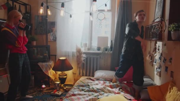若いです屈託のない男ダンスオンベッドでレトロな部屋装飾されたガーランドとして彼のガールフレンド撮影ビデオとともにヴィンテージカメラ — ストック動画