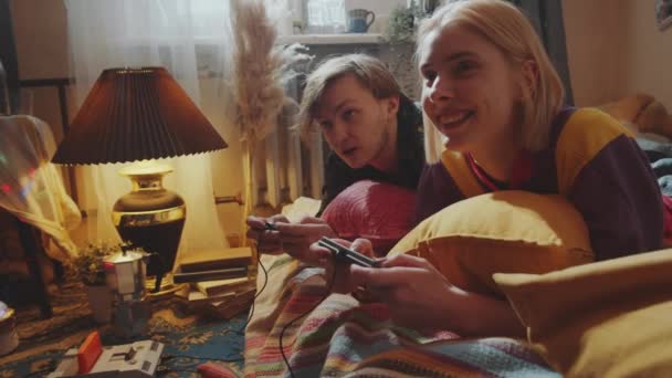 若いです白人カップル笑顔とチャットながら一緒にベッドの上に家に横たわって レトロコンソールゲームをプレイ — ストック動画