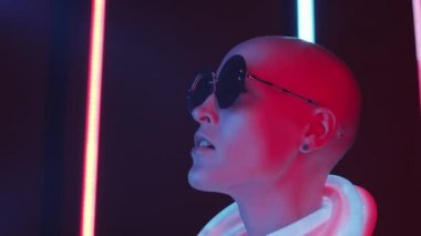 Arka planda çok renkli floresan lambalarla karanlık stüdyoda poz verirken şık yuvarlak güneş gözlüklü ve parlak neon tüplü kel mankenin göğüs yukarı çekimi.