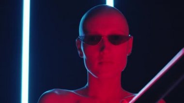 Şık güneş gözlüklü çıplak kadın model ve karanlık stüdyoda neon ışıklı kamera için poz veriyor.