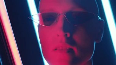 Gözlüklü, parlak bir lamba tutan ve karanlık stüdyoda poz verirken kameraya bakan kel bir kadının yakın çekimi.