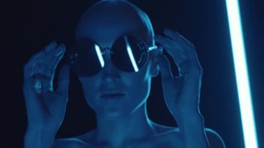 Genç kel bir kadının son moda güneş gözlüklerini çıkartıp karanlık stüdyoda çok renkli neon ışıklarla poz verirken çekilmiş el kamerası görüntüleri.