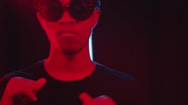 Moda gözlüklü genç Afrikalı Amerikalı adamın, arka planda yanıp sönen neon ışıkları ve floresan lambalarıyla karanlık stüdyoda gülümseyip dans ederken çekimini kaldır.