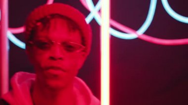 Dar güneş gözlüklü, bereli, kapüşonlu, gülümseyen ve arka planda parlak tüplerle karanlık stüdyoda dans eden genç hippi Afrikalı Amerikalı adamın göğüs yukarı çekimi.