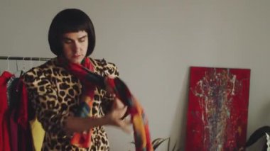 Esmer peruklu transseksüel adam atkı ve palto takıyor, aynaya bakıyor ve evi terk ediyor.