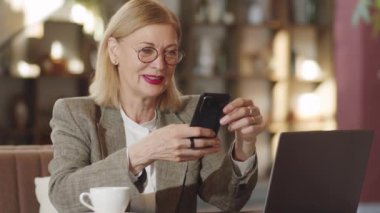 Güzel kıdemli iş kadını kafeteryada dizüstü bilgisayarı ve kahvesiyle oturuyor, akıllı telefon kullanıyor ve gülümsüyor.