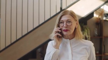 Resmi giyimli güzel bir iş kadını ofiste yürüyor ve cep telefonuyla konuşuyor.