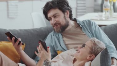 Oturma odasındaki kanepede oturan genç ve pozitif bir çift akıllı telefonlarıyla internette geziniyor ve karantinada birlikte otururken bir şeyler hakkında sohbet ediyorlar.