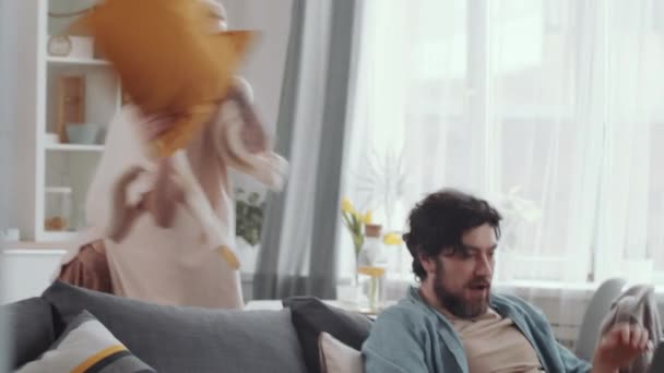 Bearded Caucasian man sitting on couch and working on laptop at home. Joyous wife hitting him with pillow and laughing, then playful couple having pillow fight in the living room