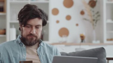 Sakallı beyaz adamın kredi kartı tutarken ve dizüstü bilgisayar kullanırken evde kanepede otururken ve online alışveriş yaparken videoyu indirirsin.