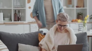 Kanepede dizüstü bilgisayar kullanan neşeli beyaz kadın kocasını arıyor. Neşeli çift tartışıyorlar, online dükkandan sipariş veriyorlar ve kredi kartıyla ödüyorlar.