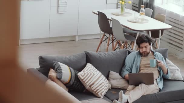 一个大胡子白人男子坐在舒适的沙发上 一边喝着杯子里的茶 一边在笔记本电脑上浏览互联网 一边在网上购物 拍了一张高视角的照片 — 图库视频影像
