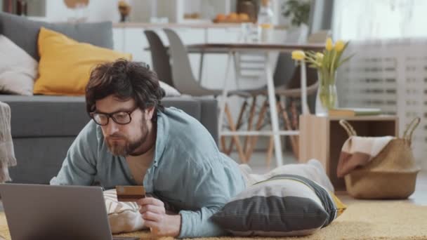 留胡子的白种人 戴着眼镜躺在客厅的地板上 在笔记本电脑上输入信用卡的细节 同时在家里的网店购物 — 图库视频影像