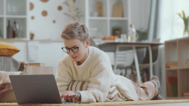 年轻快乐的女人 戴着眼镜躺在客厅的地板上 手里拿着信用卡 一边在网上购物 一边在家里自我封闭 — 图库视频影像