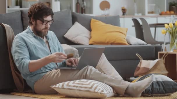 在家里 戴眼镜 头戴胡子的白人男子坐在客厅的沙发旁边 一边在笔记本电脑上输入信用卡号码 一边在网上商店购买商品 — 图库视频影像
