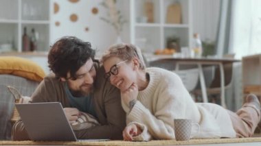 Mutlu aile çifti oturma odasında yerde birlikte yatar, internette dizüstü bilgisayara bakar ve karantina sırasında kameraya bakar ve gülümserler.