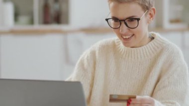 Güzel sarışın kadının seçici odak fotoğrafını çek mutfak masasında otururken, dizüstü bilgisayarına kredi kartı numarası yazarken çevrimiçi dükkandan mal alırken, gülümseyip fincandan çay içerken.