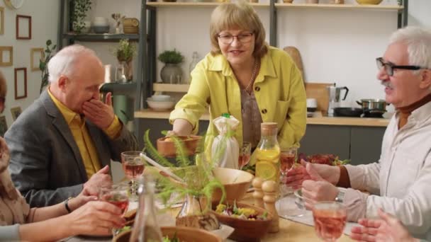 一位快乐的老妇人在家里主持节日晚宴时 一边把菜递给朋友 一边坐在桌旁 — 图库视频影像