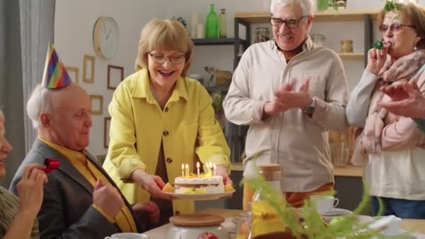 戴着派对礼帽的快乐老人坐在餐桌旁 在生日蛋糕上吹着蜡烛 一边微笑一边拍手 一边和朋友们一起庆祝 — 图库视频影像