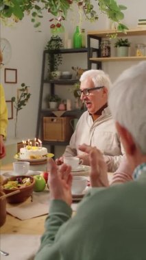 Doğum günü pastasında mum üfleyen ve arkadaşları yemek masasında el çırparken karısını öpen yaşlı adamın dikey çekimi.