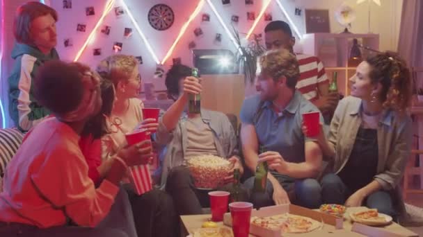 乾杯する若い白人男性 ネオンライトで飾られたリビングルームでパーティーを持っている間 多民族の友人や飲み物ビールのカップとボトルをクリック — ストック動画