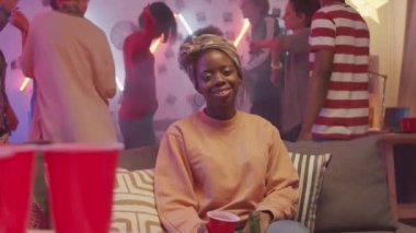 Genç, güzel, Afro-Amerikalı bir kadın kanepede oturup kameraya gülümserken arkadaşları arka planda dans ediyor.