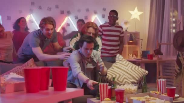 喜びの友人が彼を引っ張っながら ビールのボトルとソファに座って退屈男は ネオンで飾られた部屋のパーティーで一緒に踊る — ストック動画