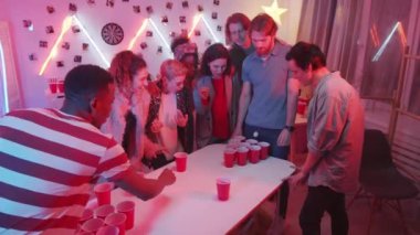 Bir grup heyecanlı arkadaş, havalı ev partisinde bira pong oyununda kaybederken içki içip masaya bardak fırlatan genç beyaz adam için tezahürat yapıyorlar.