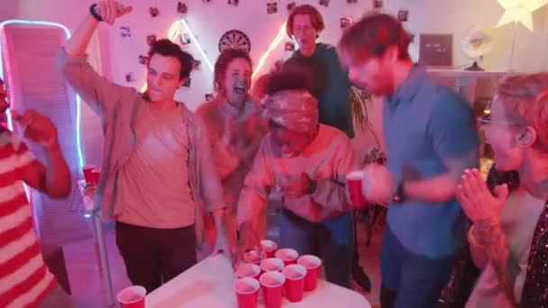 若い陽気なアフリカ系アメリカ人の女性が赤いカップから飲んで 友達とホームパーティーでビールポンポンゲームをしている間に腕を上げて笑顔を上げる — ストック動画