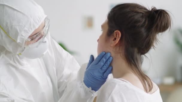 女性患者の首にリンパ節を検査しながら自宅でコロナウイルス検査を行う防護服 マスクの女性医師 — ストック動画
