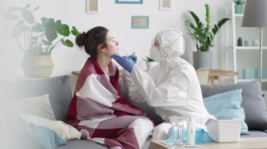Koruyucu üniformalı bir sağlık görevlisi kanepede genç bir kadınla oturuyor, burnundan örnek alıyor ve evde Coronavirus testi yaparken virüs taşıyıcısına örnek koyuyor.