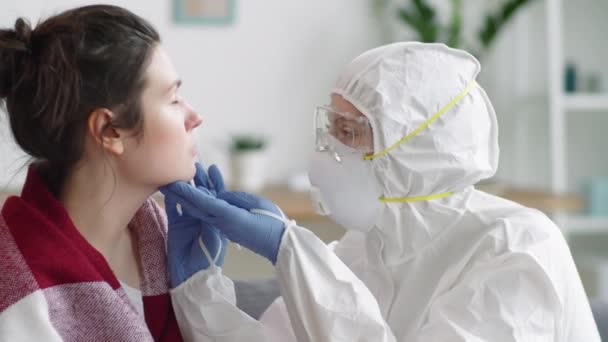 女性医師で保護スーツ 眼鏡と顔マスク収集鼻スワブから若い女性と話して彼女にしながら 彼女のためにウイルスのために彼女をテスト自宅 — ストック動画