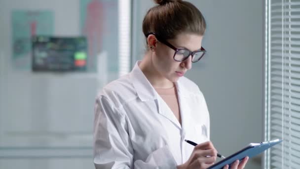 身穿实验室外套和眼镜的年轻专业女医生站在窗边写着便条的剪贴板上 诊所里戴着百叶窗 — 图库视频影像