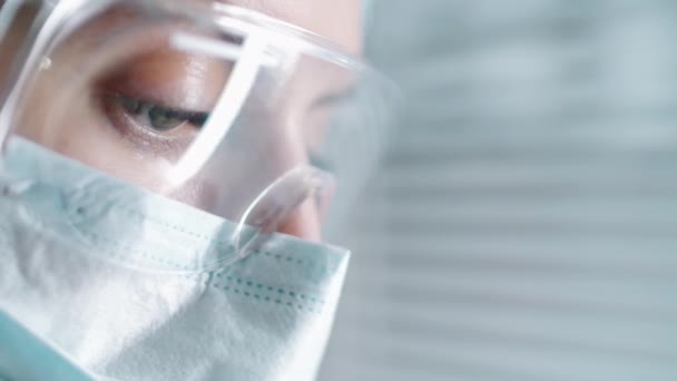 病院の窓の近くに立っている間 保護眼鏡やフェイスマスクで女性医師の顔のショットを閉じて 医学文書を見下ろし 読んでください Covid 19流行概念 — ストック動画