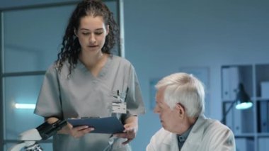 Genç bayan asistan, üst düzey erkek bilim adamına belgelerle birlikte pano getiriyor ve laboratuarda birlikte çalışırken onunla konuşuyor.