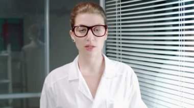 Laboratuvar önlüklü ve gözlüklü genç beyaz kadın doktor hastanede kameraya bakıyor ve bir şey hakkında konuşuyor.