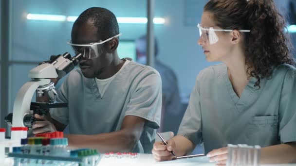 Young Black Male Scientist Uniform Looking Microscope Talking Caucasian Female — Stock Video