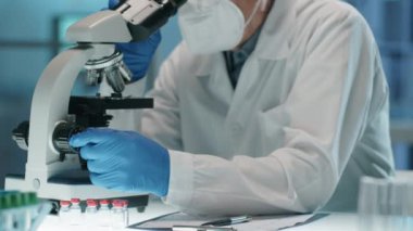 Laboratuvarda çalışırken koruyucu eldivenler, maske, gözlük ve steril şapka takan yaşlı bilim adamının mikroskopla görüntüsünü yukarı kaldır.