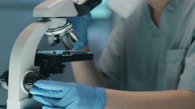 Koruyucu eldivenli, maskeli, gözlüklü ve şapkalı kadın bilim adamının mikroskopla bakarken laboratuvarda araştırma yaparken çekimini kaldır.