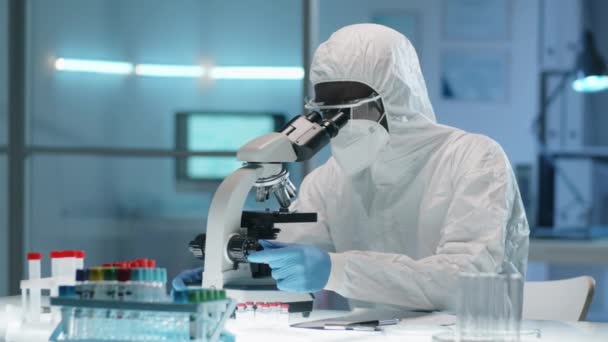アフリカ系アメリカ人の男性科学者は 保護服 マスク 眼鏡を顕微鏡で観察し その後 研究室の机でノートを取りながら コイド中に働く パンデミック — ストック動画