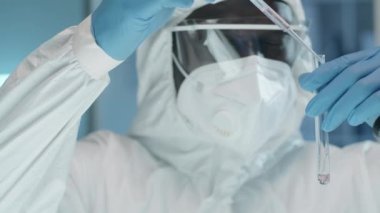 Korunma giysisi, maske, eldiven ve gözlüklü Afro-Amerikan erkek bilim adamının koronavirüs salgını sırasında laboratuvarda kimyasal deney yaparken pipetle test tüpüne sıvı dökerken yakın plan çekimi.