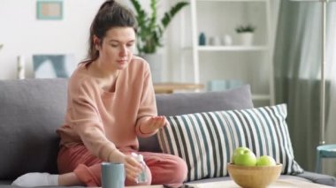 Genç bir kadın dezenfektanla ellerini ovalıyor ve evdeki kanepede otururken akıllı telefona uyguluyor. Coronavirus önleme kavramı