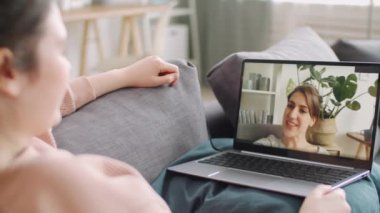 Genç bir kadın dizüstü bilgisayarıyla kanepede dinleniyor ve izole bir şekilde evde otururken neşeli bayan arkadaşıyla sohbet ediyor.