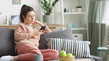 Evdeki kanepede oturup internette sörf yaparken akıllı telefondan kayan esmer saçlı beyaz bir kadının orta boy görüntüsü.