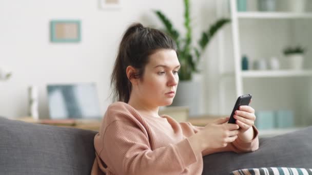 若いですブルネット女性座っていますソファに自宅とスクロールソーシャルメディア上のスマートフォン — ストック動画