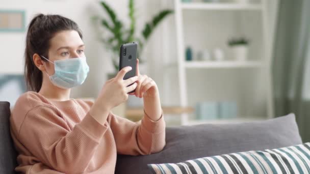 リビングでソファに座っている保護医療用マスクの若い女性とロックダウン中に自宅に滞在しながらスマートフォンでソーシャルメディアをスクロール — ストック動画