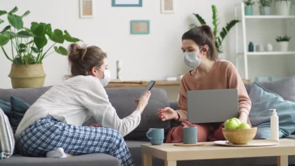 两名戴着医疗面罩的年轻女性坐在客厅的沙发上 一边在智能手机和笔记本电脑上浏览互联网 一边呆在家里休息 — 图库视频影像